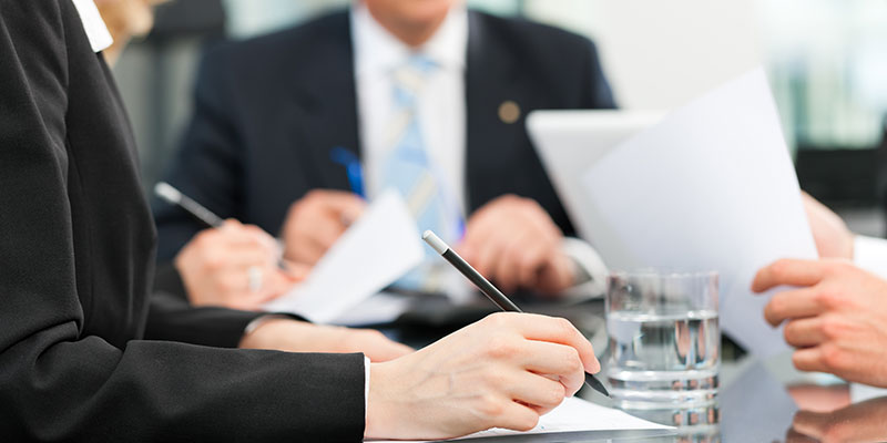 group of people dressed in business attire around a table having a discussion and taking notes and passing around paper
